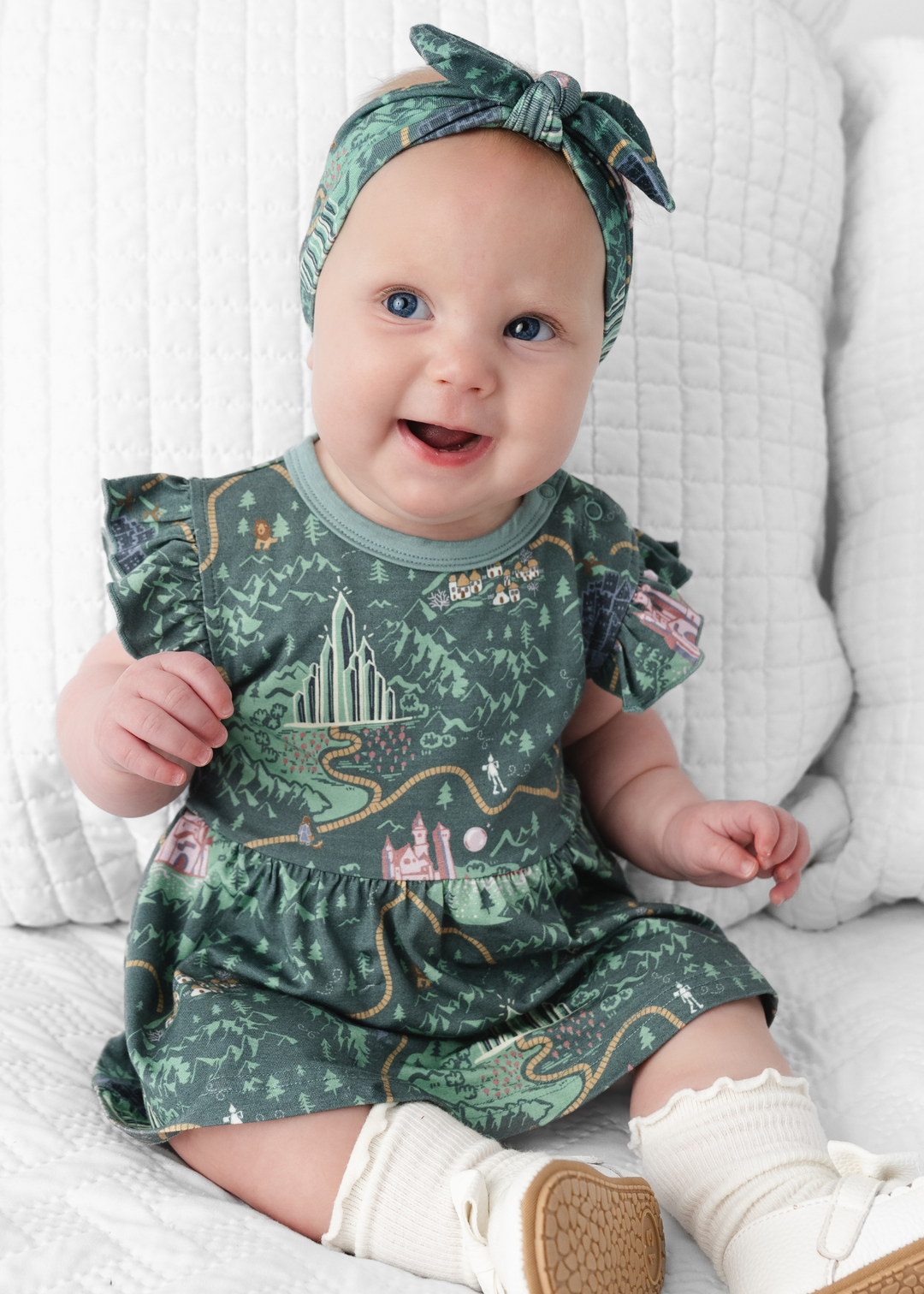 A baby wearing a green flutter sleeve dress with a Wizard of Oz pattern and a matching headband.