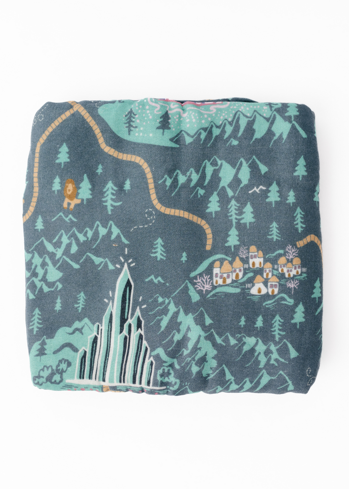 A swaddle blanket with a vibrant green tone featuring a whimsical pattern with characters and a landscape inspired by the Emerald City and the yellow brick road.