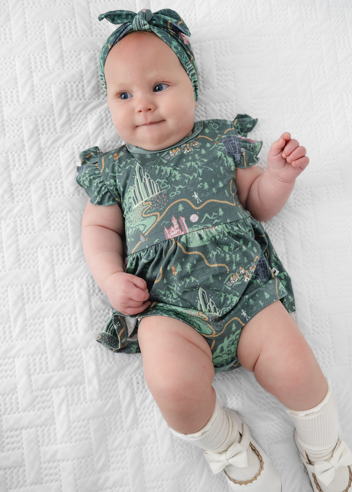 Baby girl in a Wizard of Oz short sleeve baby dress with Wizard of Oz characters, a yellow brick road and a vibrant green background. 