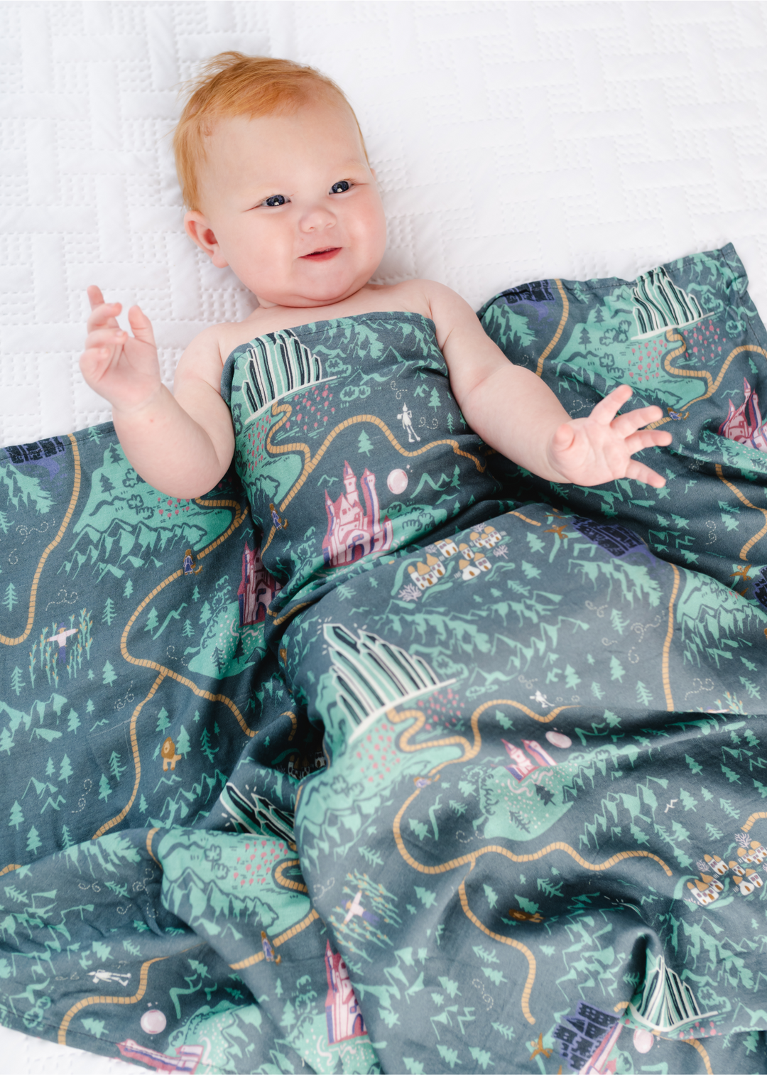 A swaddle with a green background that features a yellow brick road and several Wizard of Oz characters. 