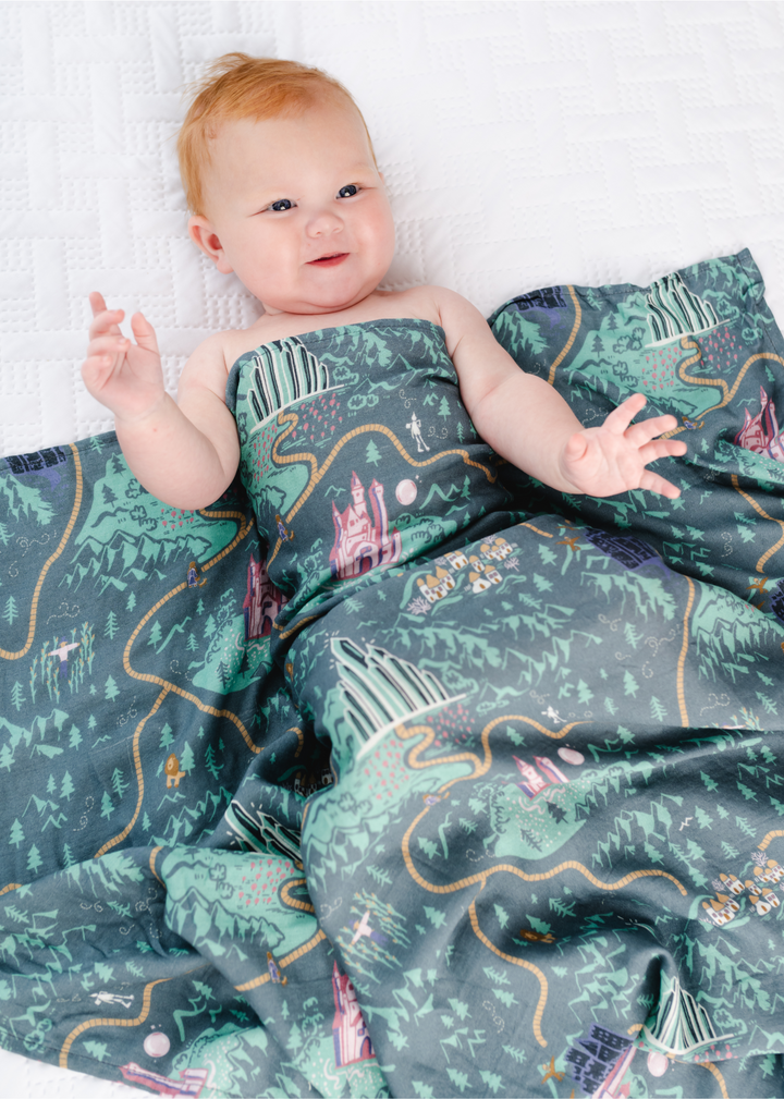 A swaddle with a green background that features a yellow brick road and several Wizard of Oz characters. 