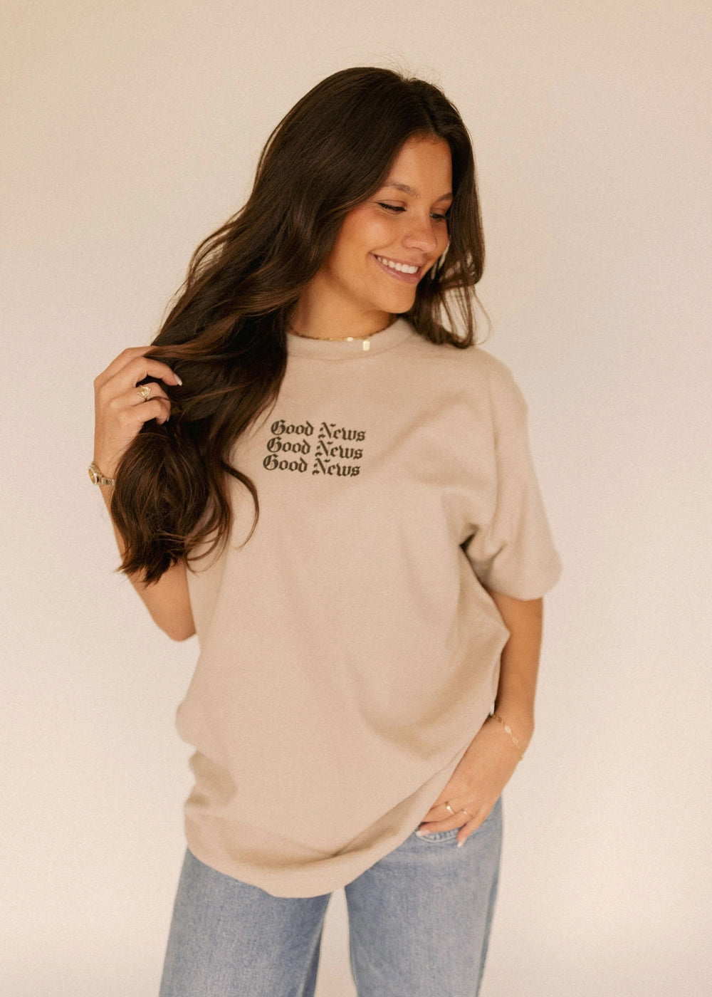 Woman wearing a beige t-shirt with Christian text "Good News", standing against a plain background