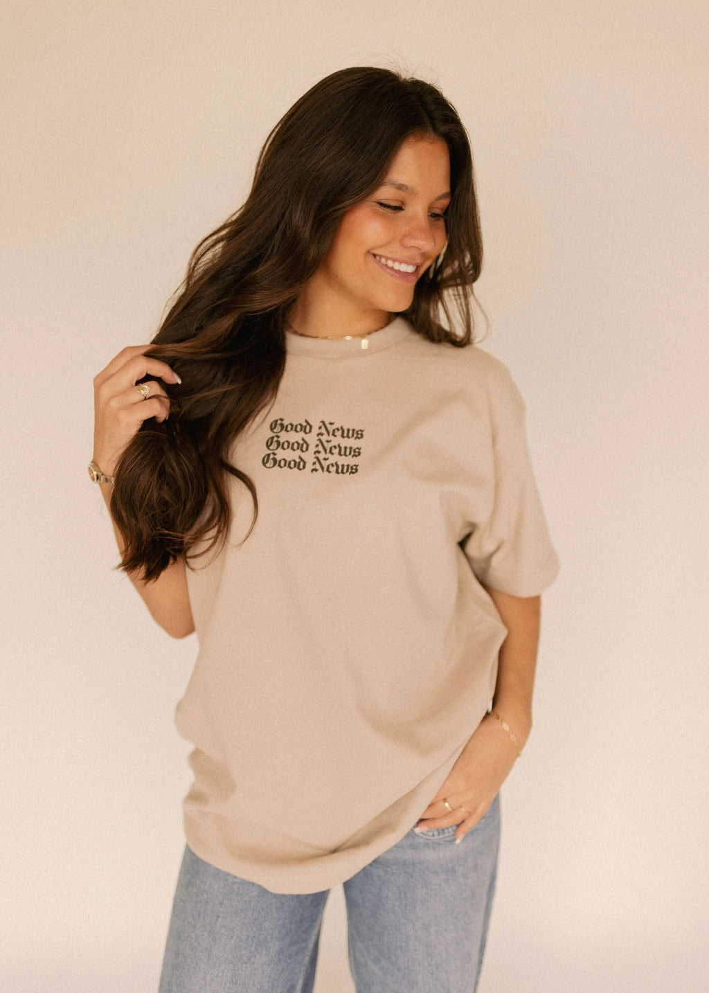 Woman wearing a beige t-shirt with Christian text "Good News", standing against a plain background