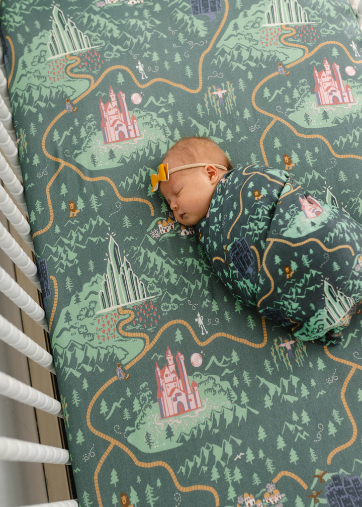 Wizard of Oz inspired matching swaddle and crib sheet on a green background with Wizard of Oz characters and a yellow brick road featured. Emerald City Bamboo Collection